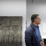 Brian, an injured workers in blue jacket, stands in front of injured worker sculpture