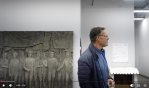 Brian, an injured workers in blue jacket, stands in front of injured worker sculpture